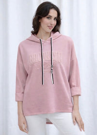 A woman wearing a dusty pink pullover hoodie with 'BROOKLYN' and 'New York' embroidered on the front, paired with white pants, standing against a white curtain backdrop.