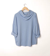 A light blue draped cowl-neck top with rolled three-quarter sleeves, hanging on a wooden hanger against a plain white wall.