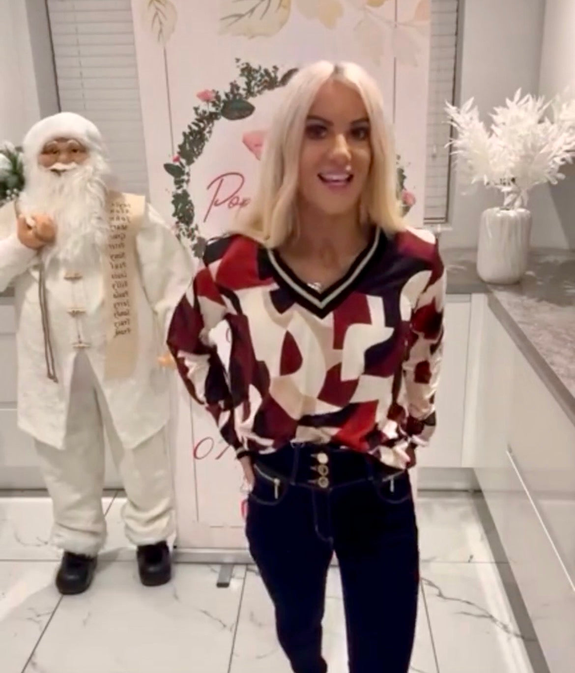Woman in a Cindy Metallic V-Neck Print Top standing next to a person in a Santa Claus costume in a decorated room.
