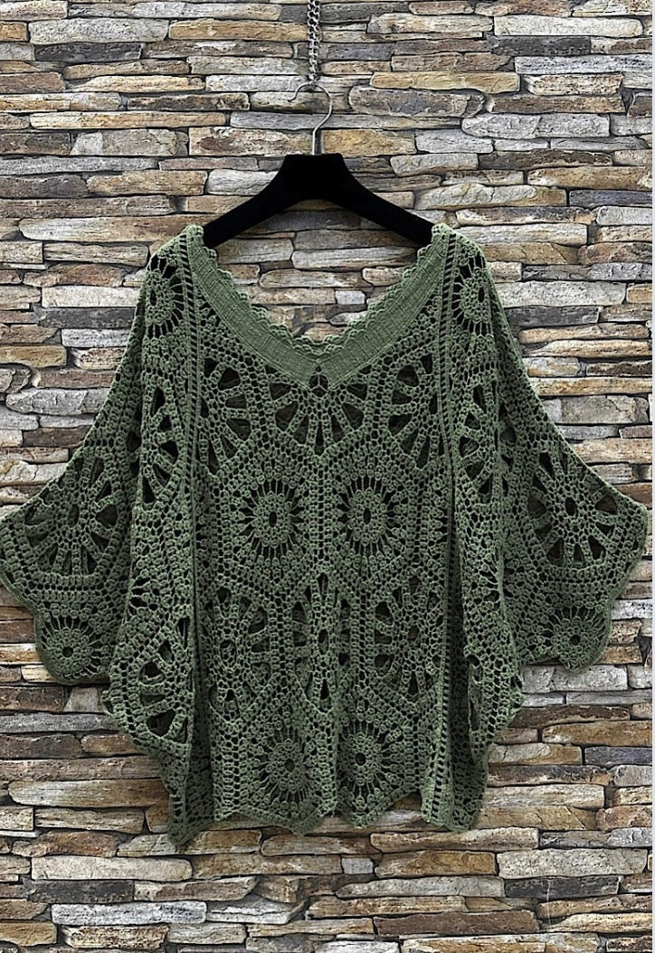 Green Drew Jumper on a hanger against a stone wall background