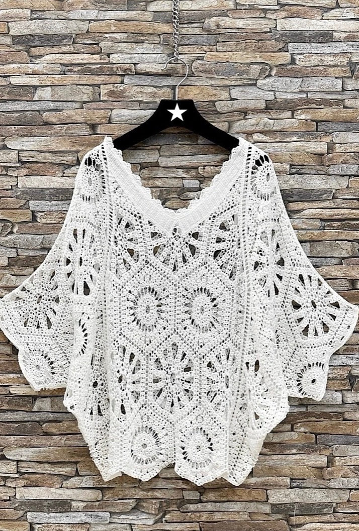 White crochet poncho on a hanger against a stone wall background