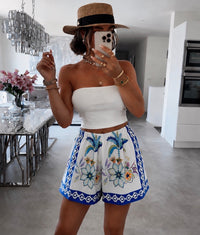 Person taking a mirror selfie wearing a white top and floral embroidered shorts in a modern kitchen.
