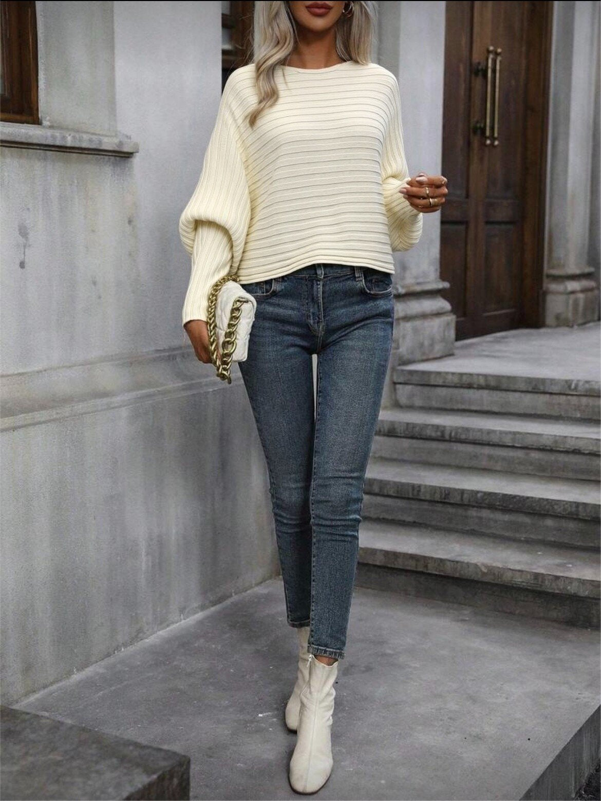 Person wearing a cream sweater, blue jeans, and white boots standing on steps. "Emily Batwing Top"