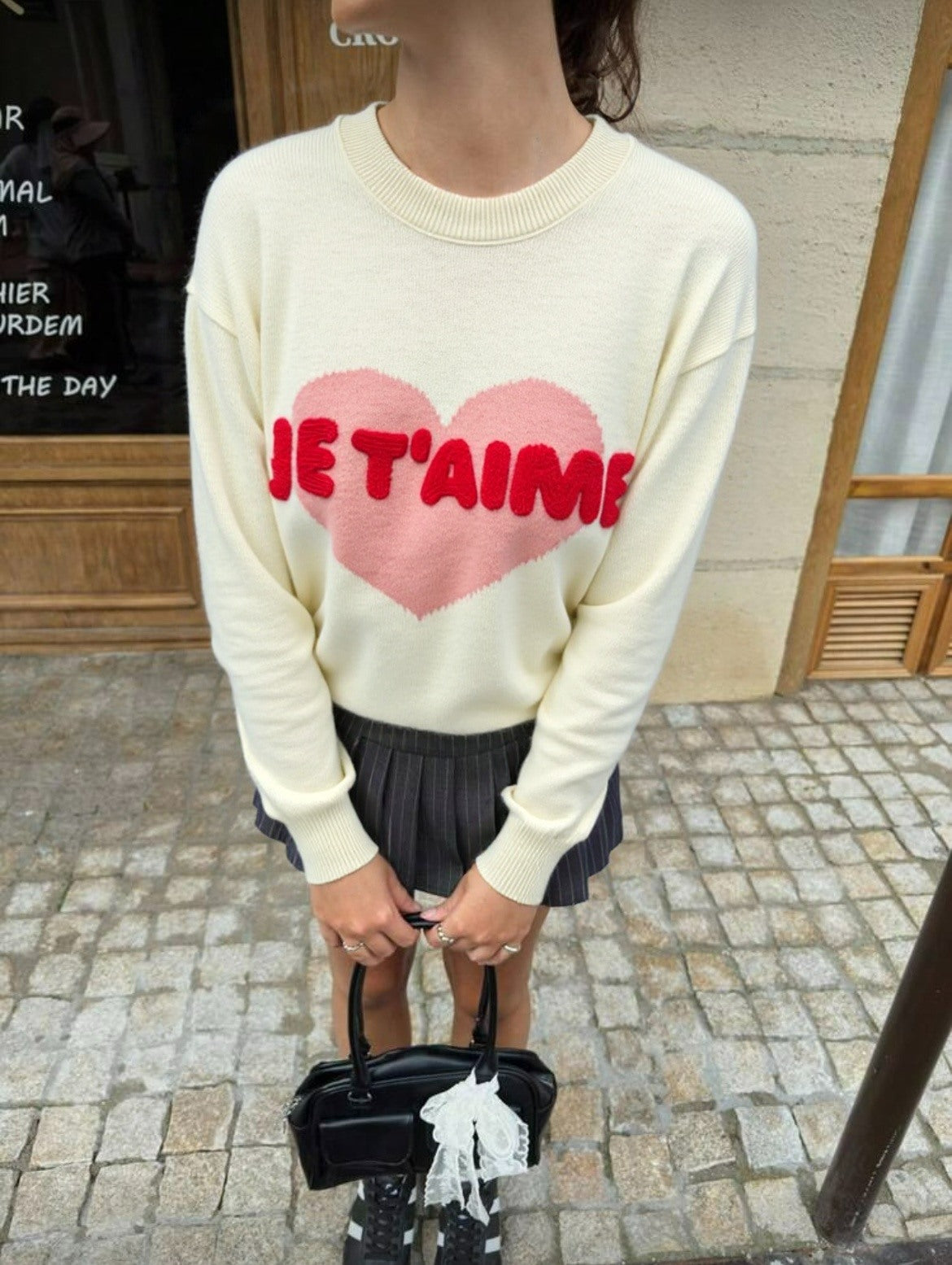 Person wearing a cream sweater with a pink heart and red text, standing on a street. "Emily Knit Comfortable Everyday Knitwear"