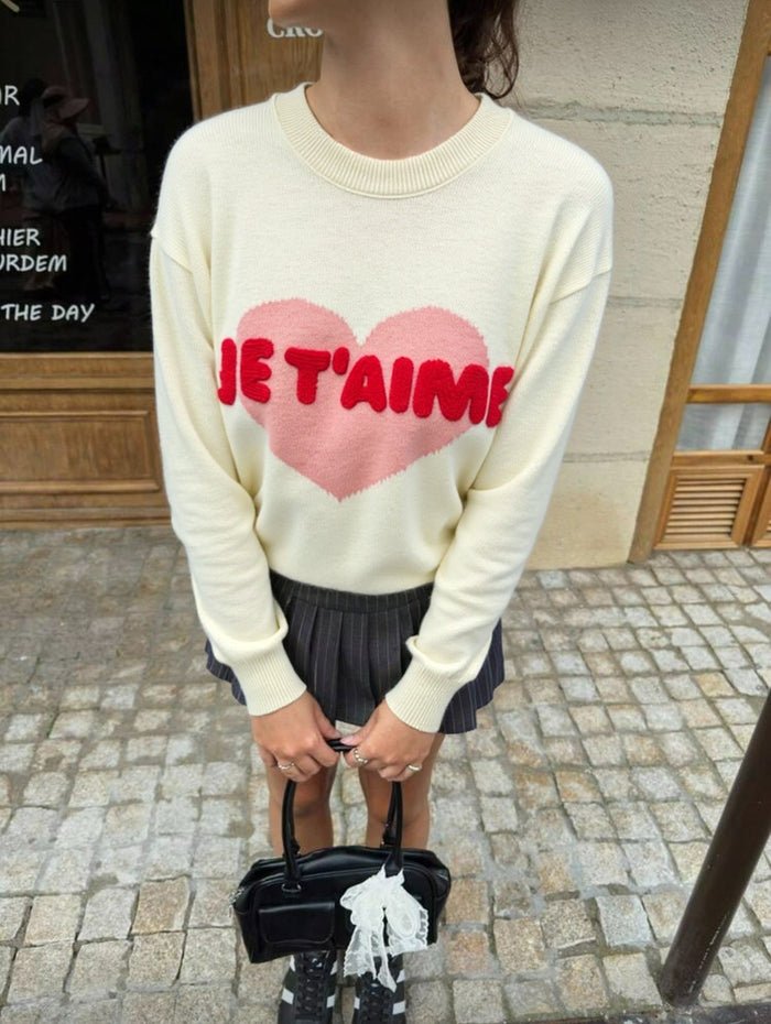 Person wearing a cream sweater with a pink heart and red text, standing on a street. "Emily Knit Comfortable Everyday Knitwear"