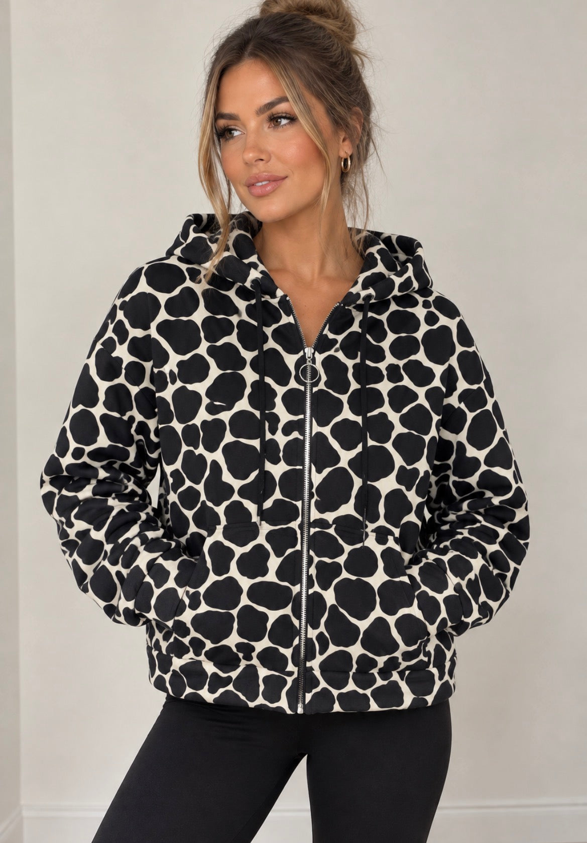 A woman wearing a black and off-white cow-print zip-up hoodie jacket with a front zipper, hood, and side pockets, paired with black pants, set against a plain light gray wall.