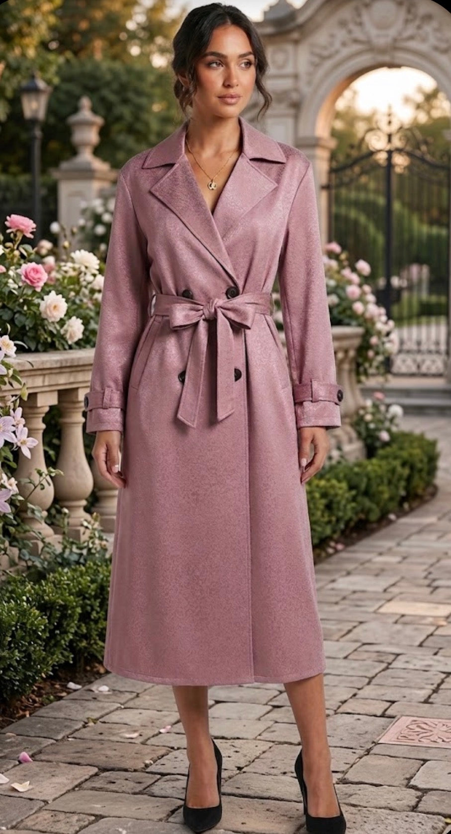 A woman models a dusty rose double-breasted trench coat with a matching waist tie, standing on a stone pathway in a garden setting.
