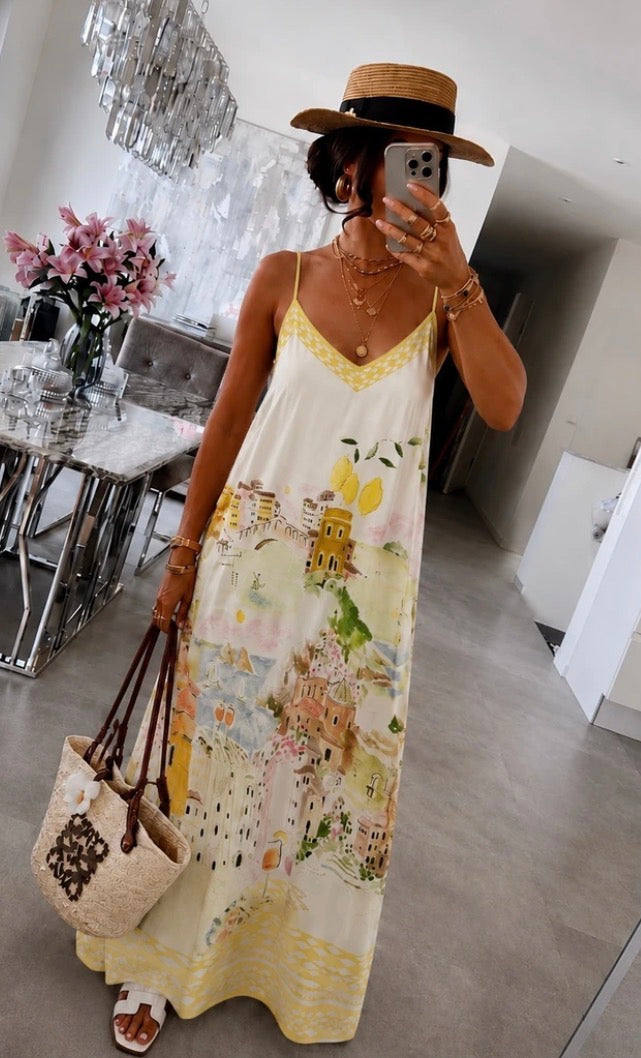 A woman takes a mirror selfie wearing a long cream maxi dress with a yellow floral and botanical print, a straw hat, and carrying a woven tote bag in a modern indoor setting.