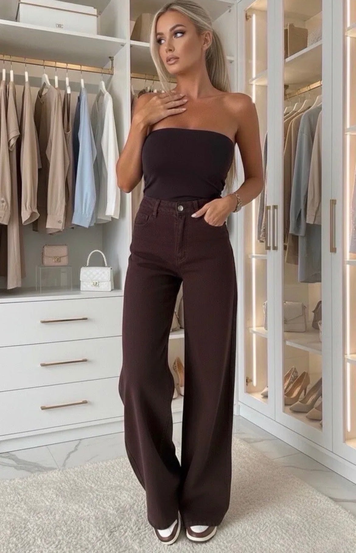 A woman stands in a bright, modern walk-in closet wearing a dark brown strapless bandeau top and matching dark brown wide-leg pants.