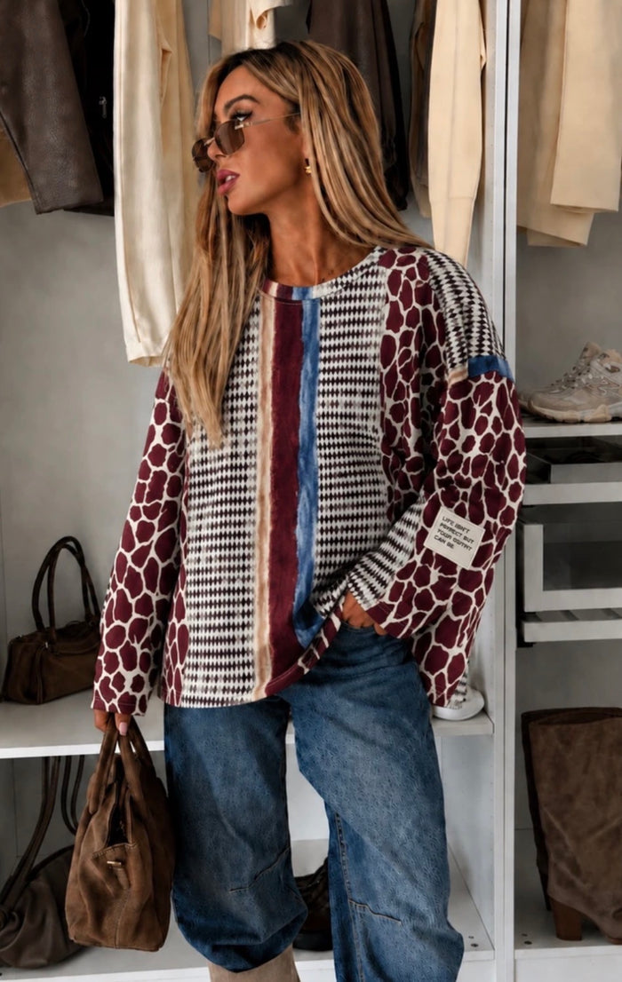 A woman models a long-sleeve patchwork knit top with a houndstooth pattern, vertical stripes, and giraffe print panels, paired with blue jeans in a closet setting.