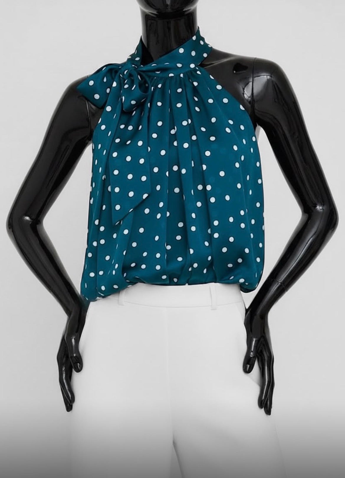 A teal halter-style blouse with white polka dots, displayed on a black mannequin torso against a light gray studio background.