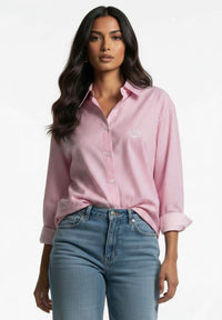 A woman modeling a pink and white vertically striped button-down shirt with a small embroidered logo on the chest, paired with blue jeans, against a plain white background.