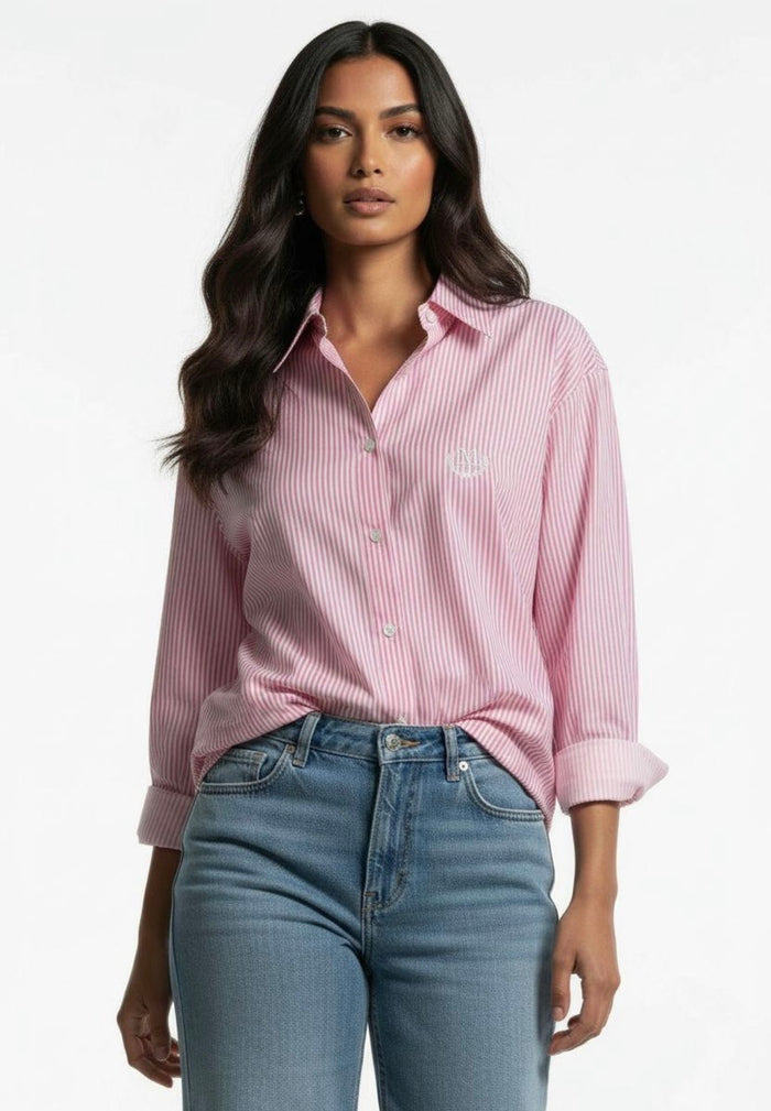 A woman modeling a pink and white vertically striped button-down shirt with a small embroidered logo on the chest, paired with blue jeans, against a plain white background.