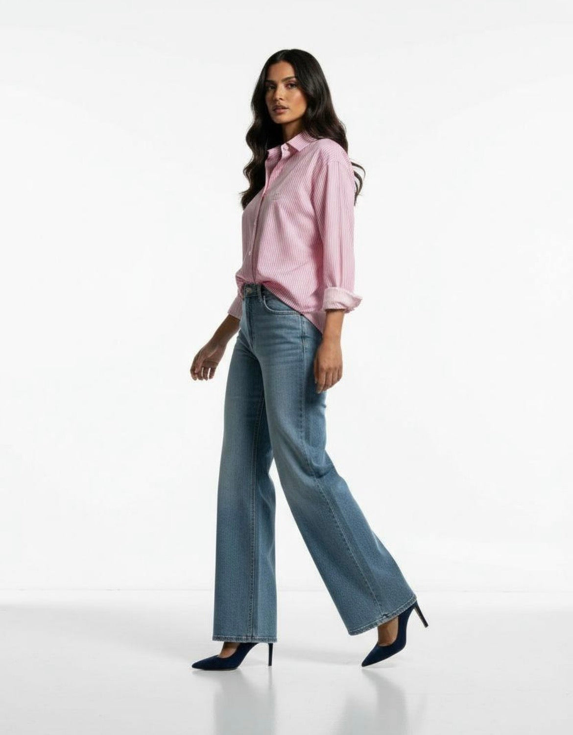 A woman modeling a light pink button-down shirt with rolled sleeves and wide-leg blue jeans, paired with dark blue high heels, against a plain white studio background.