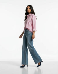 A woman modeling a light pink button-down shirt with rolled sleeves and wide-leg blue jeans, paired with dark blue high heels, against a plain white studio background.