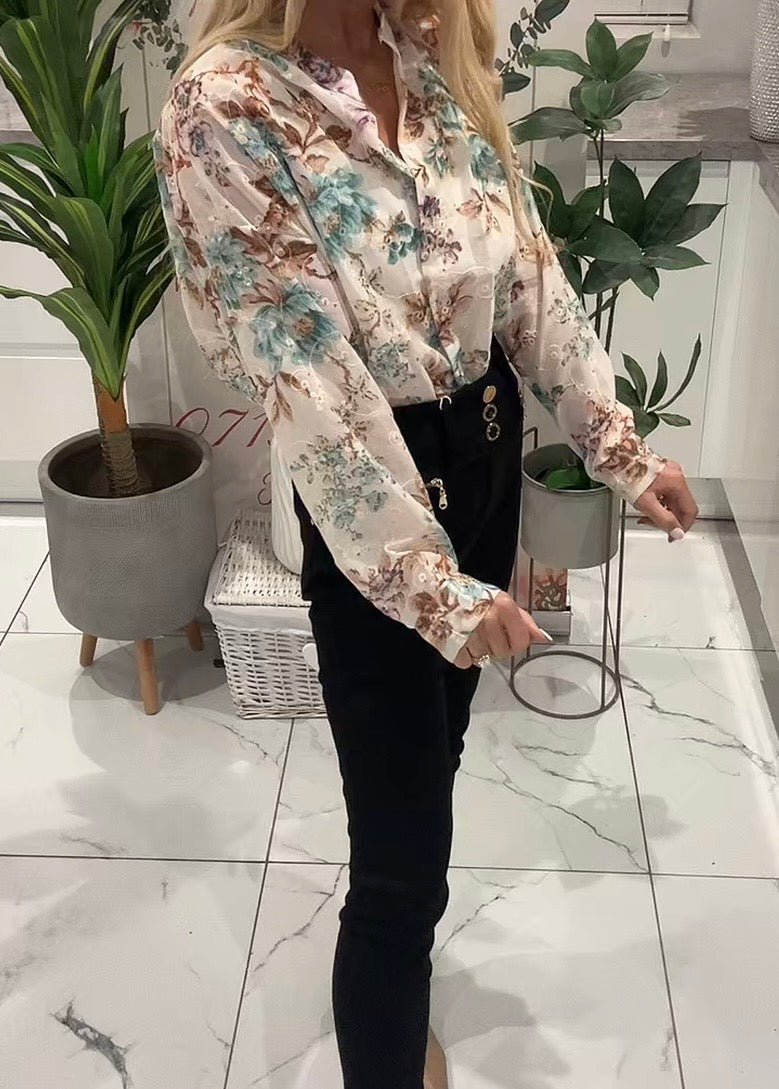 Person wearing a floral blouse and black pants in an indoor setting with plants and a basket.