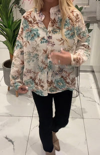 Person wearing a floral blouse and black pants in an indoor setting with plants.