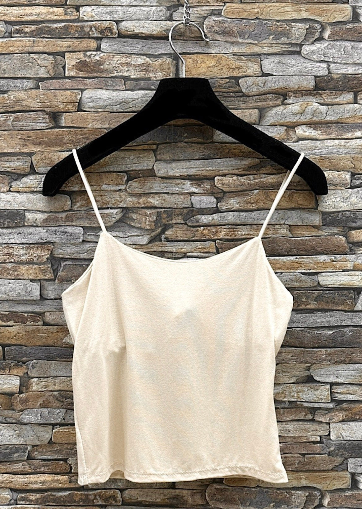Beige spaghetti strap top on a black hanger against a stone wall background