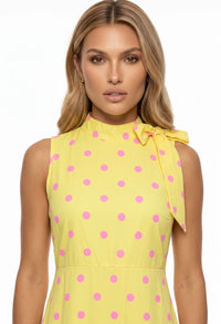 A woman wearing a sleeveless yellow dress with pink polka dots and a tie-neck detail, shown against a plain white background.