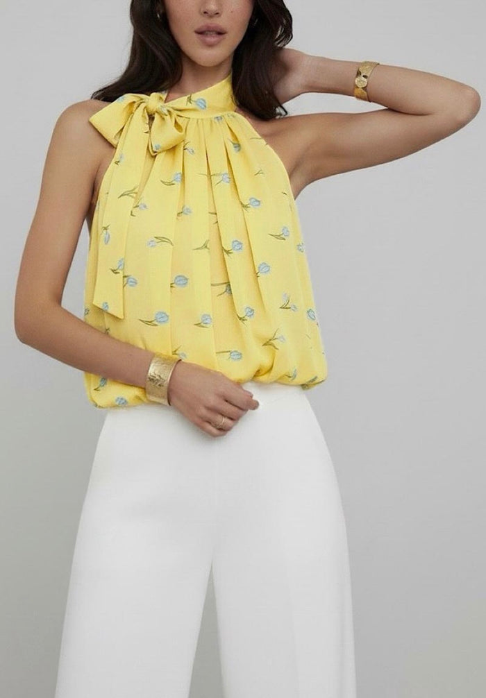 Woman wearing a yellow top with a bow detail and white pants on a plain background