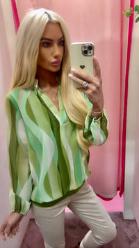 Woman taking a mirror selfie wearing a green and white patterned blouse against a pink background