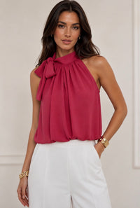 A woman wearing a sleeveless halter-style blouse with a large bow at the neck in a deep pinkish-red hue, paired with white pants, against a light neutral indoor background.