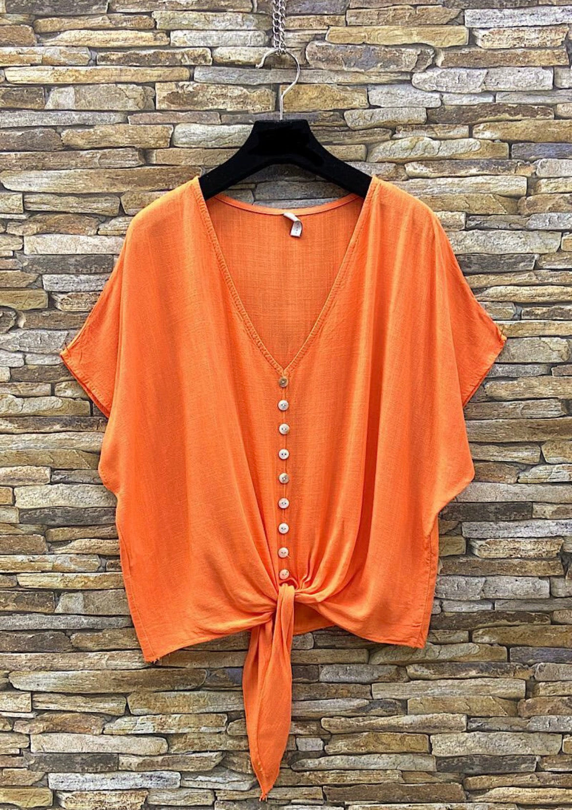 Orange top with button details on a hanger against a stone wall background