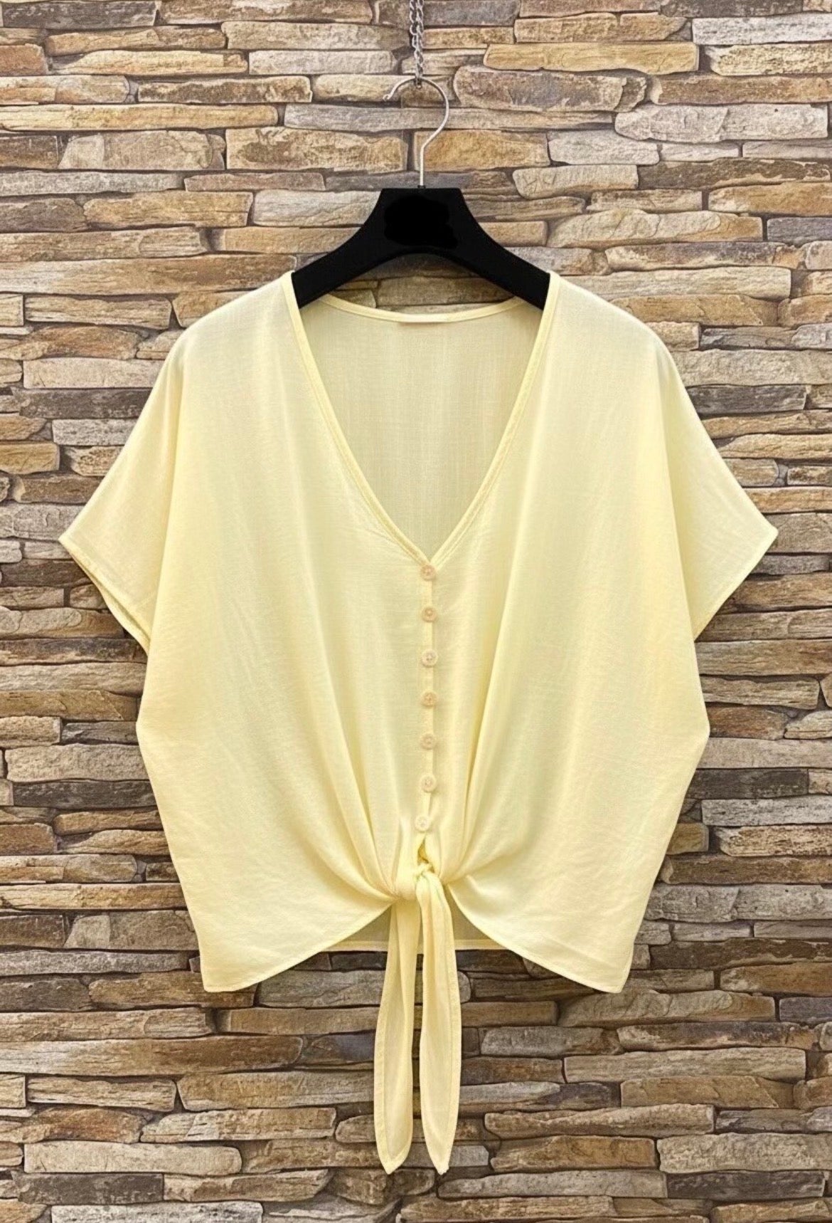 Light yellow blouse with a tie front on a hanger against a stone wall background