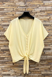 Light yellow blouse with a tie front on a hanger against a stone wall background